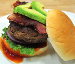 Bone Suckin Avocado Bacon Burger on plate with sauce and bun off the top, leaning on burger