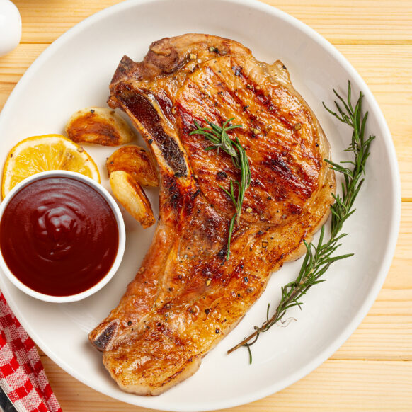 Pork chop on a plate with a cup of sauce and a sprig of rosemary